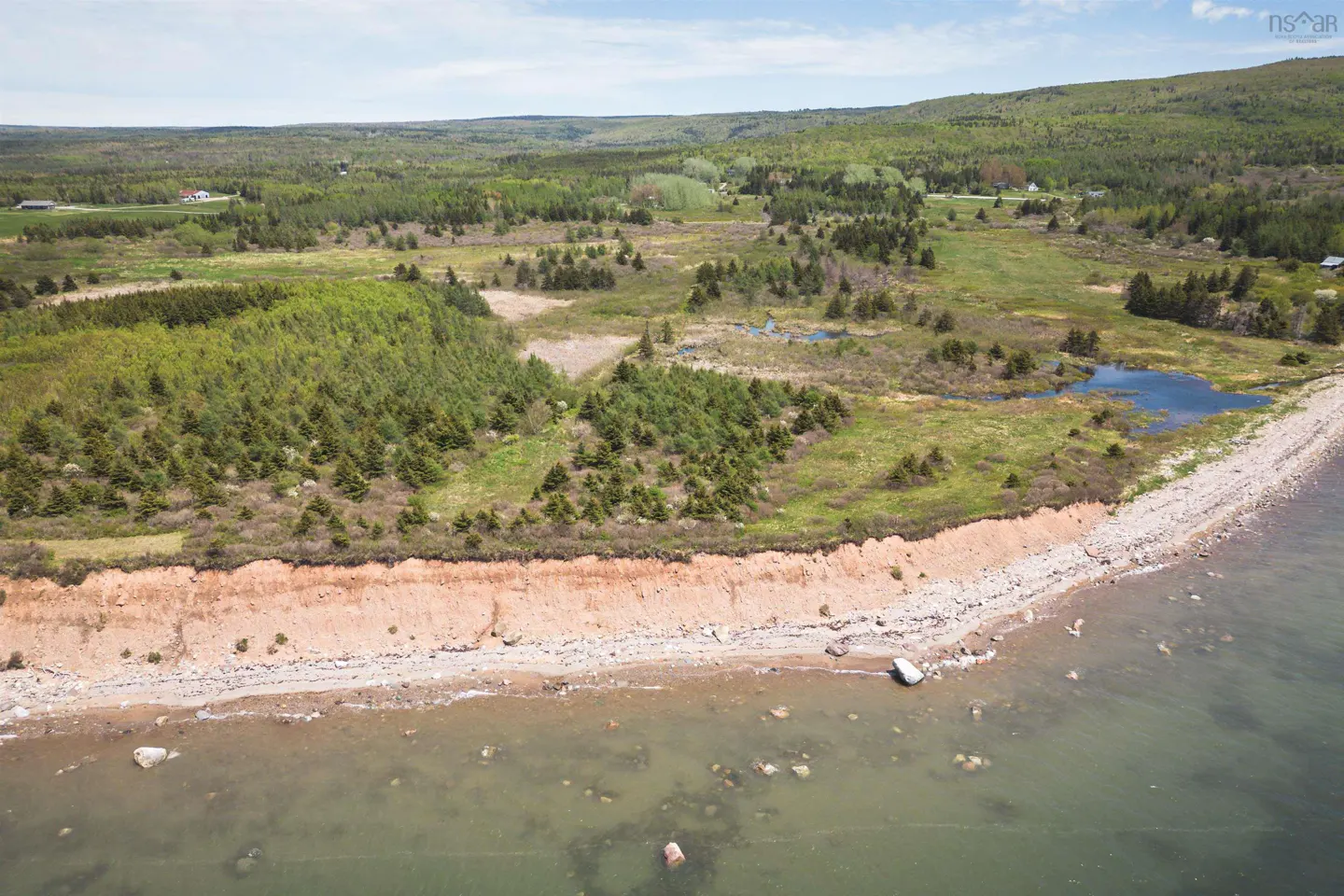 Aerial view of Nova Scotia coastline with green trees, brown cliffs, and a rocky beach meeting the clear water.