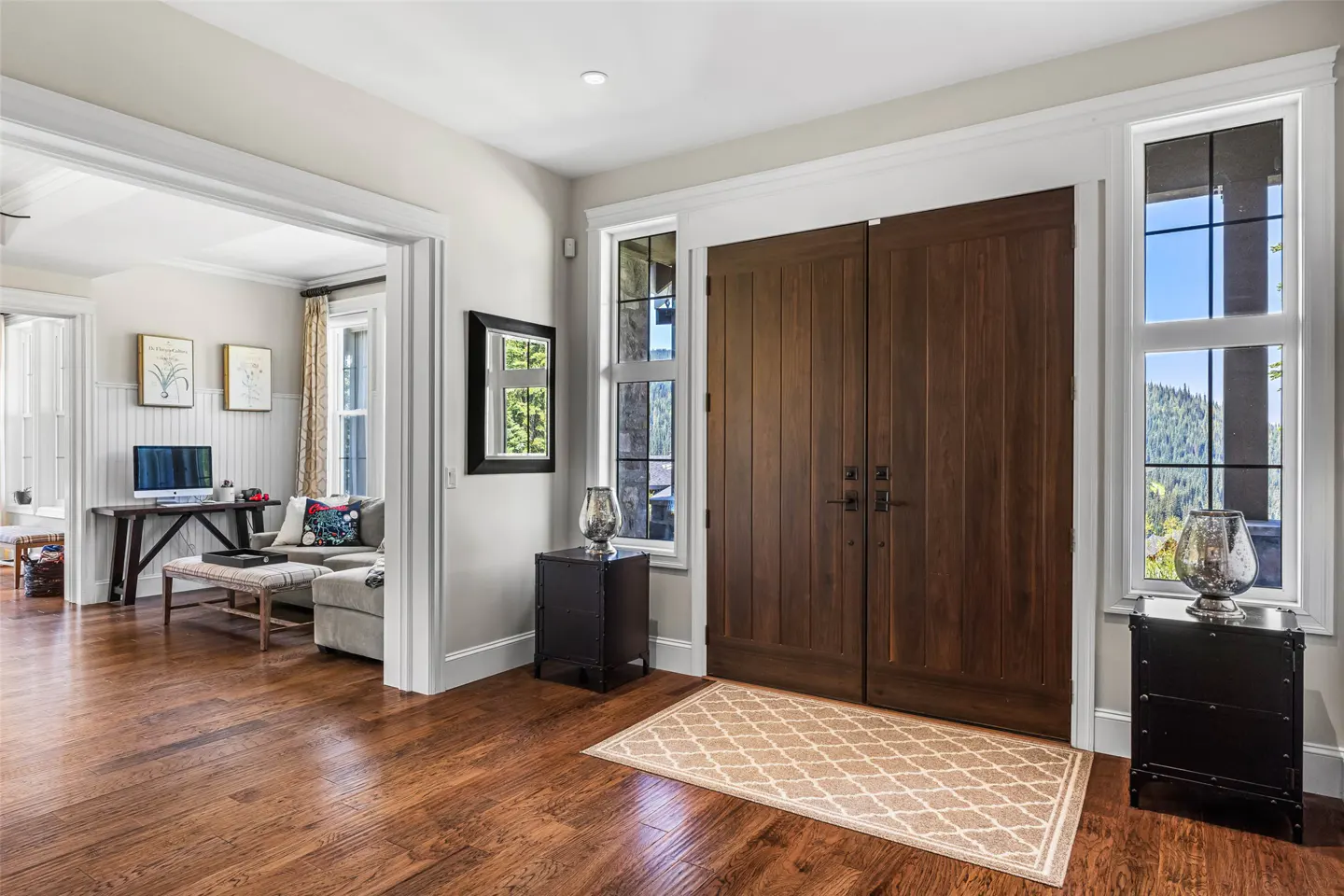 Foyer with dark wood double doors, hardwood floors, and a beige patterned rug. Windows flank the doors, offering a view of trees.