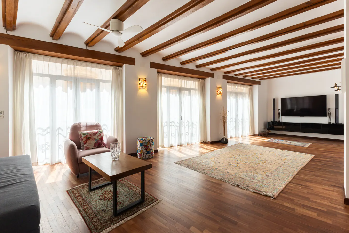 Bright living room with wood floors, exposed beams, and white walls. A large rug sits in front of a TV and entertainment center.