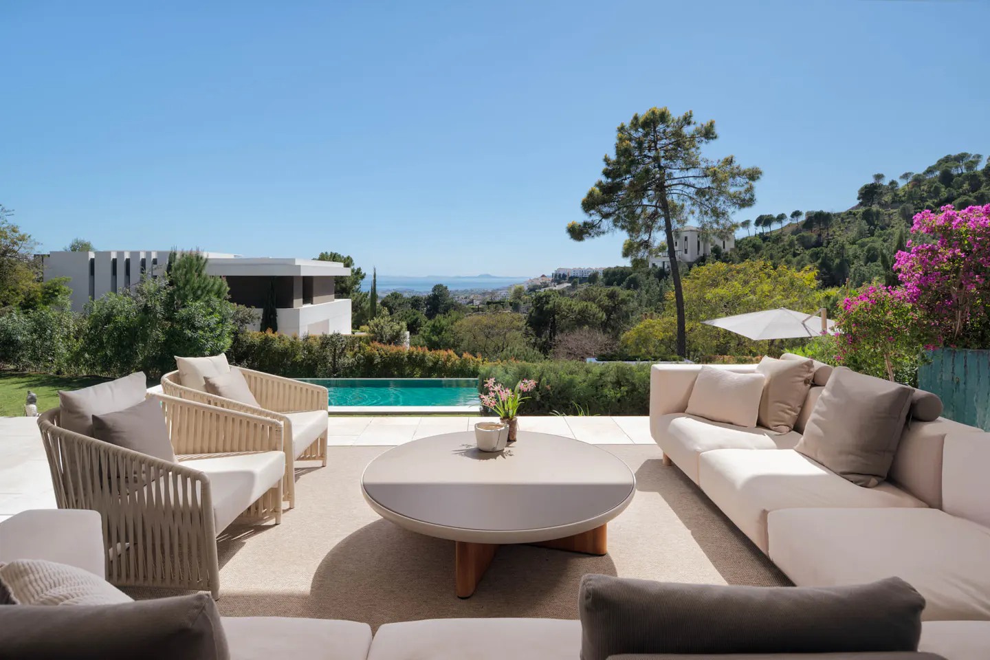Luxury patio with white sofas, rope chairs, and a round table. A pool and lush greenery are in the background under a clear blue sky.
