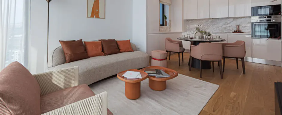 A modern apartment interior with a curved sofa, brown pillows, and orange coffee tables on a light rug. A dining area and kitchen are in the background.