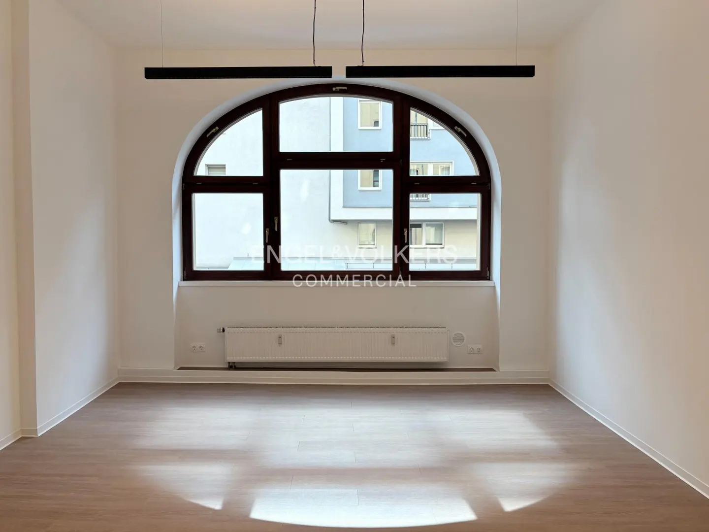 Empty room with wood floors, white walls, and a large arched window. Two black pendant lights hang from the ceiling. A radiator sits below the window.