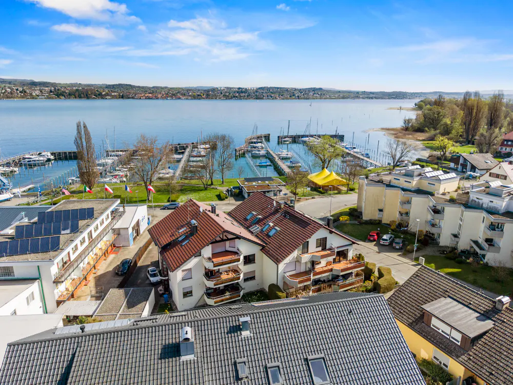 Aerial view of a marina with sailboats, surrounded by buildings with red-tiled roofs and a blue sky.