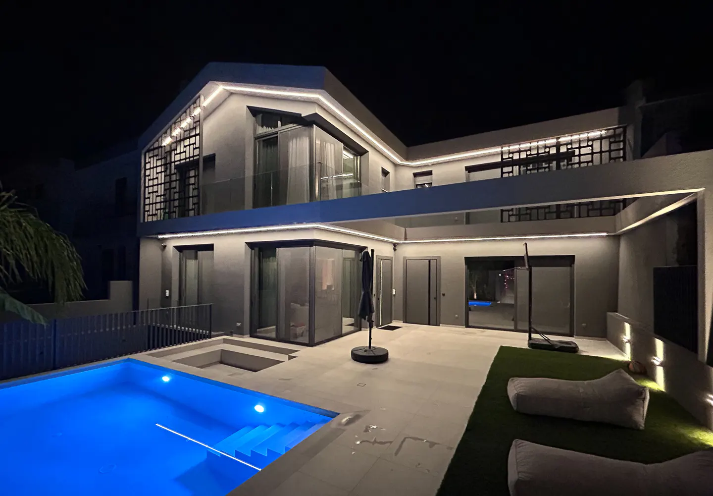 Modern two-story home at night with glowing LED trim, a bright blue swimming pool, and a patio with lounge chairs.