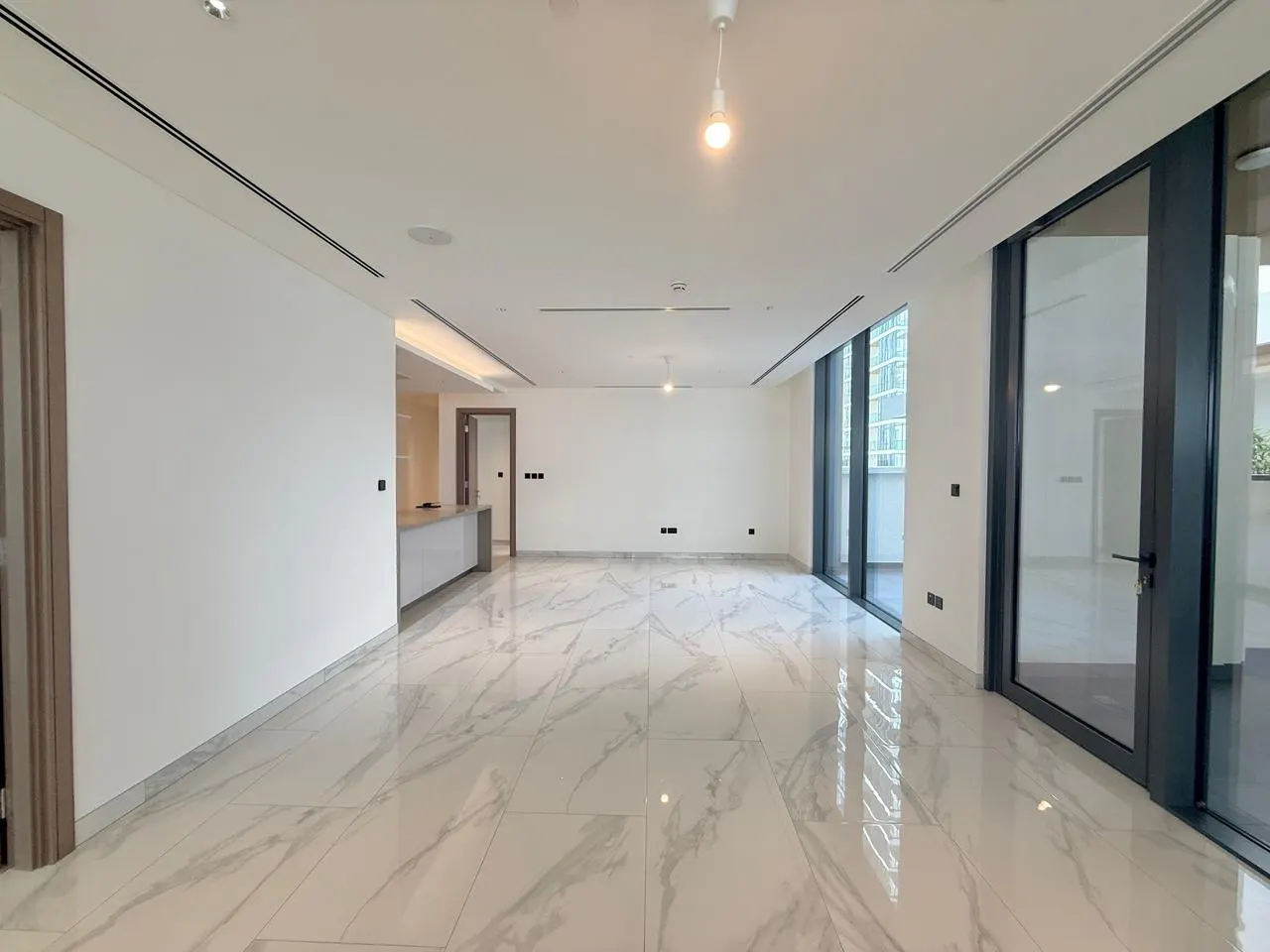Bright, empty room with white walls, marble floors, and large windows. A kitchen counter is visible in the background.