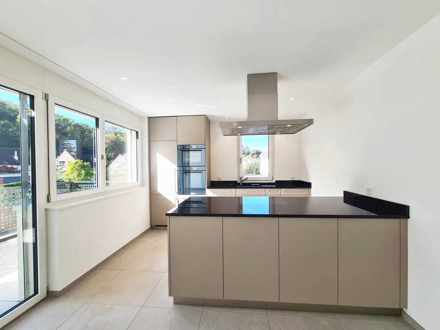 Bright, modern kitchen with beige cabinets, black countertops, and stainless steel appliances. Large windows offer a view of trees and houses.