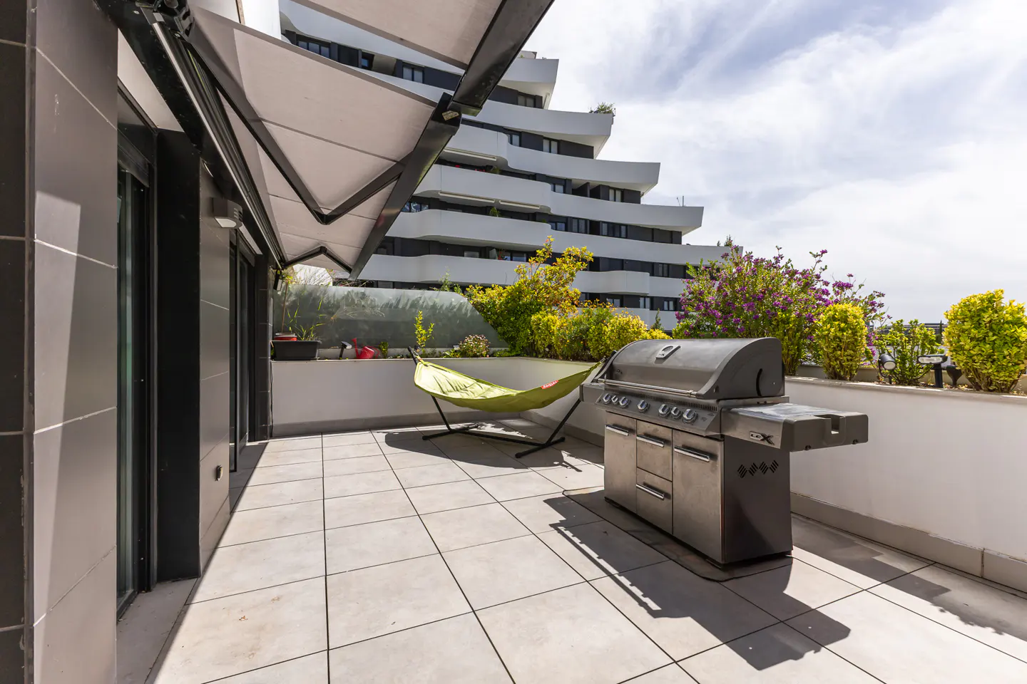 A balcony with a grill, hammock, and plants. A modern white building is in the background.