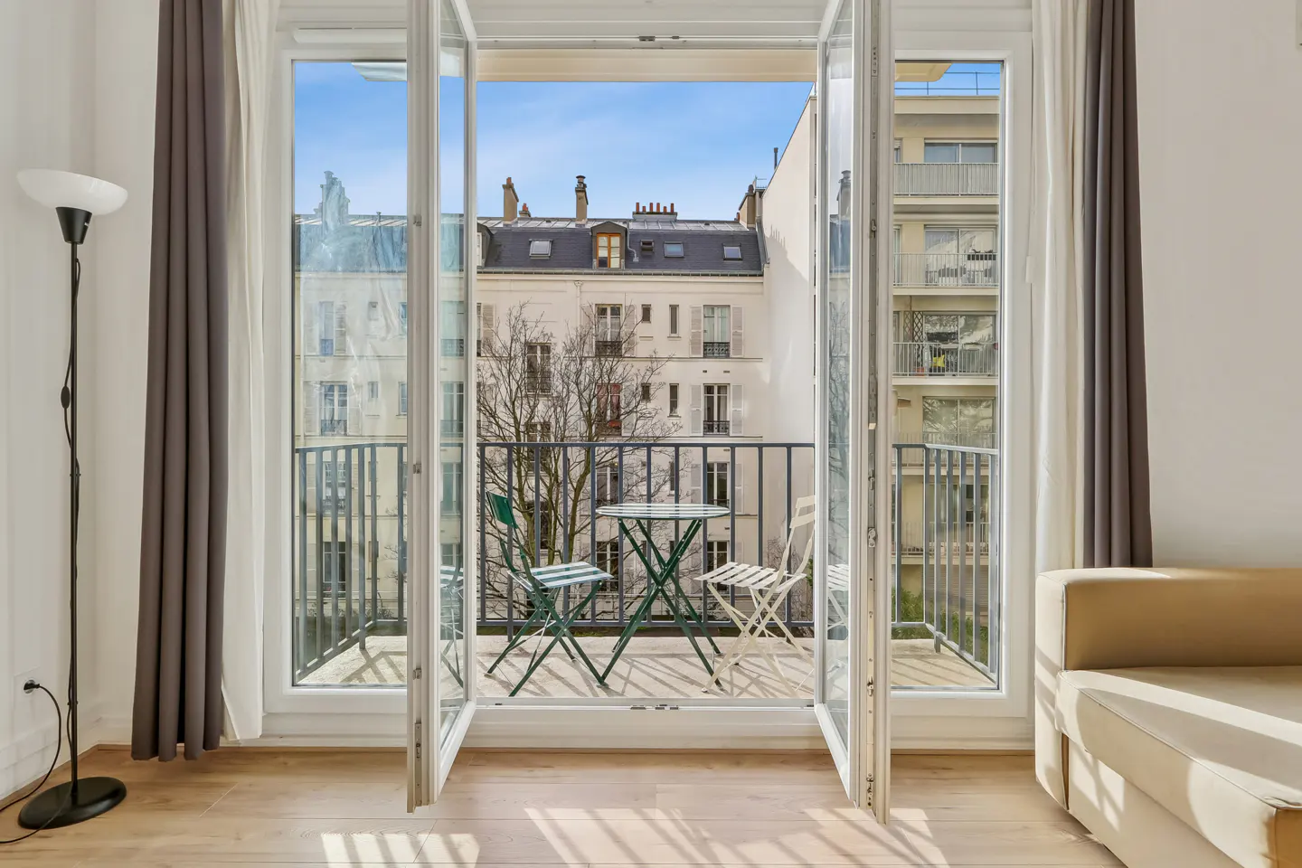 Bright apartment view through open French doors to a balcony with green table and chairs, overlooking city buildings.