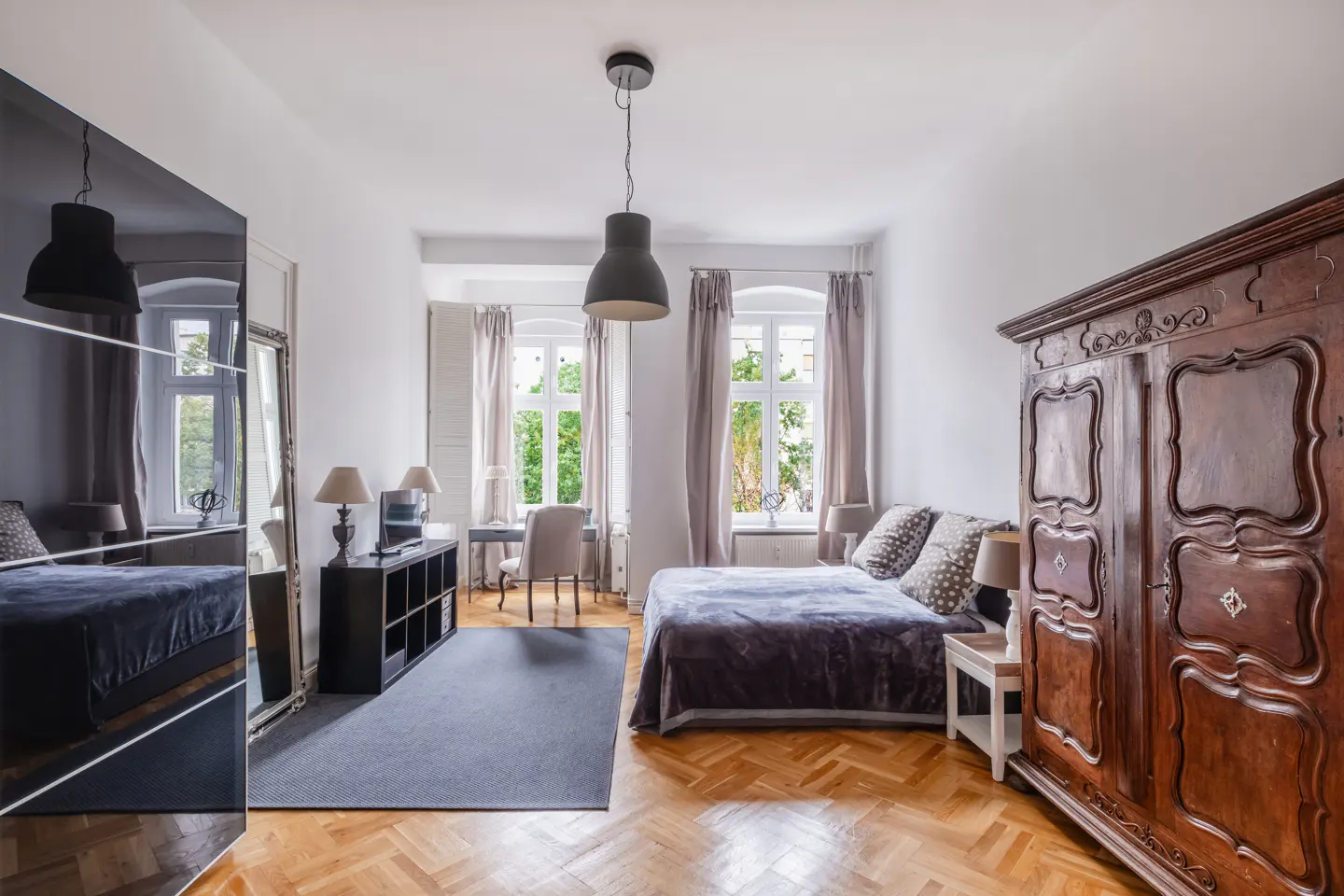 Bright bedroom with parquet floors, a bed with a gray duvet, a desk, and a large, dark wood armoire.