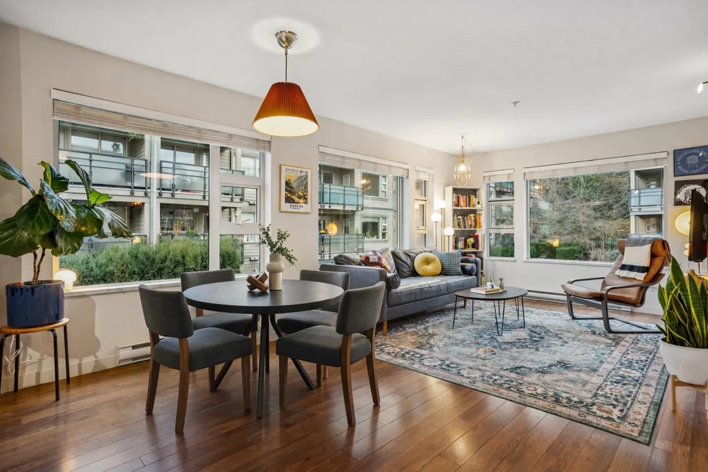 A bright living and dining area with hardwood floors, large windows, a gray sofa, and a round black dining table with gray chairs.