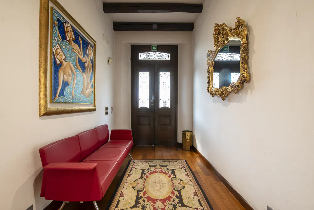 Hallway with a red sofa, ornate mirror, and a painting of women in blue. A patterned rug sits on the wooden floor, leading to a dark wood door.