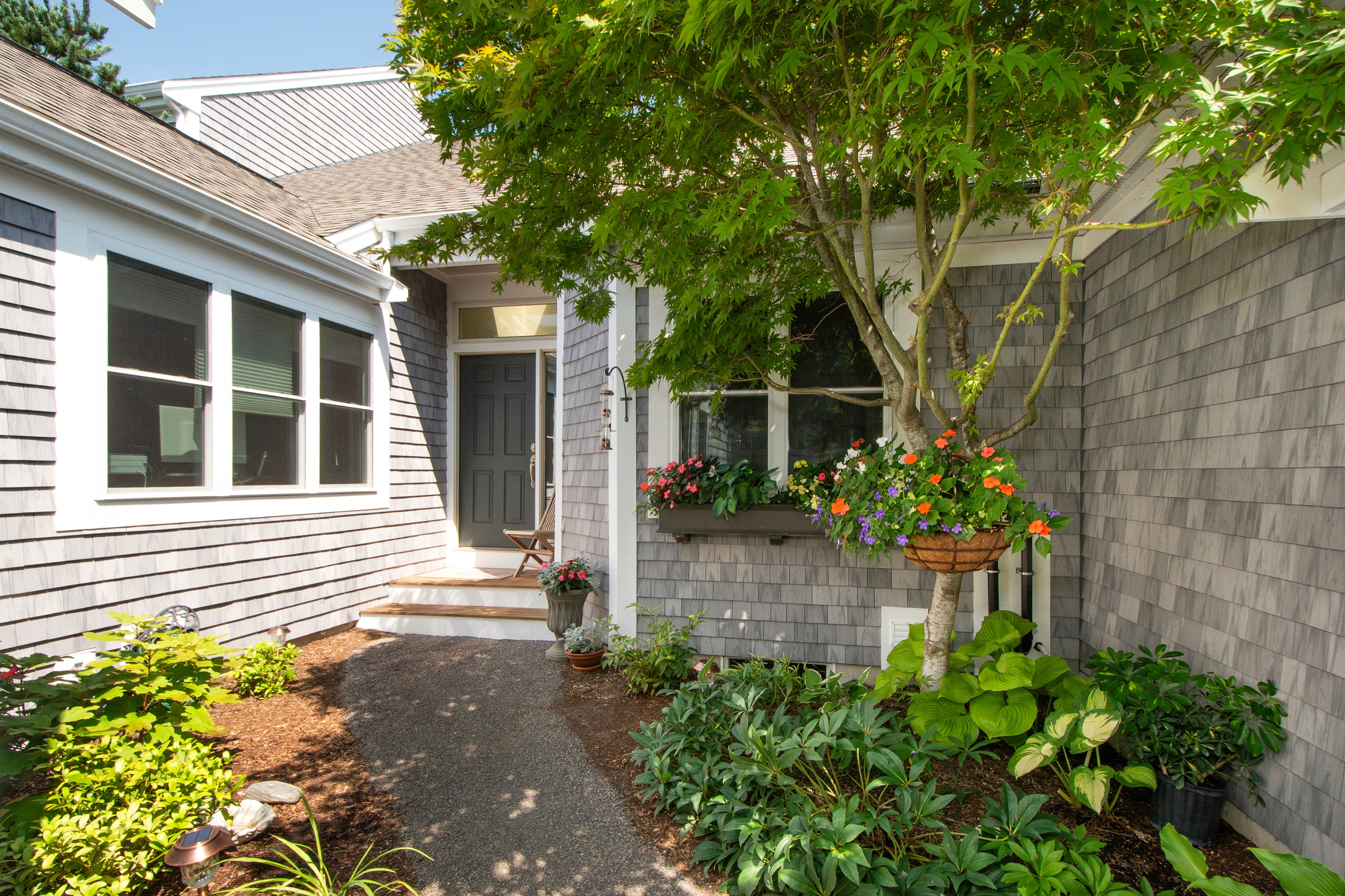 Exterior of a gray shingle home with a dark gray front door, a stone walkway, and lush green landscaping.