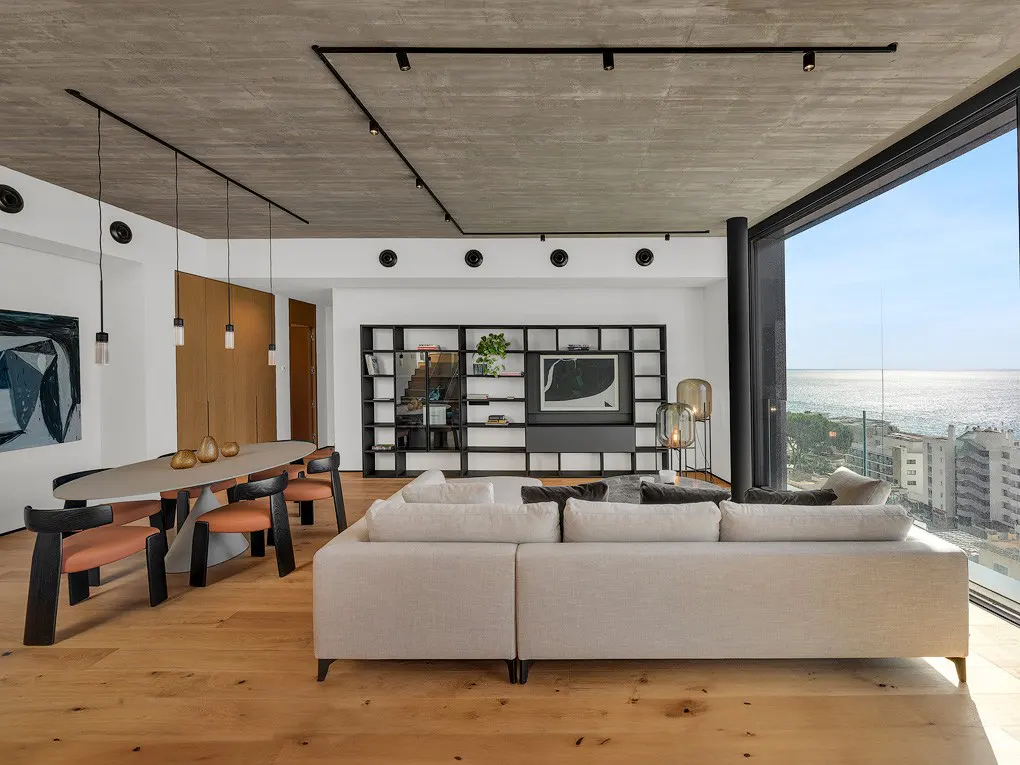 Bright, modern living room with a beige sofa, oval table with chairs, and a black bookshelf. Large windows offer a sea view.