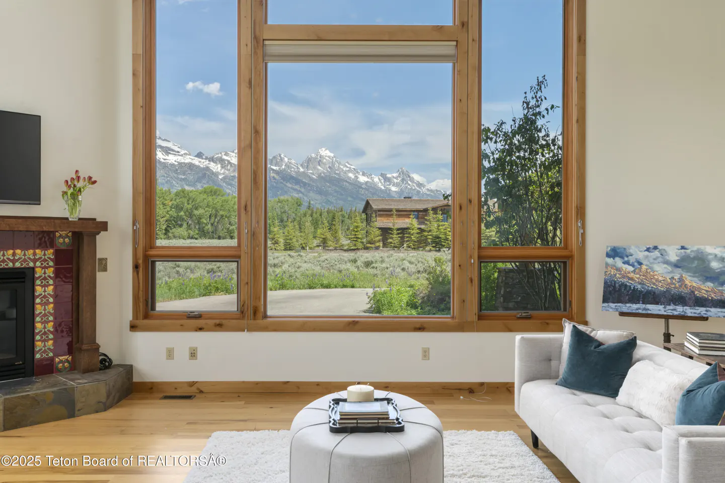 Living room with a large window view of mountains, trees, and a house. A white sofa and ottoman are in the foreground.