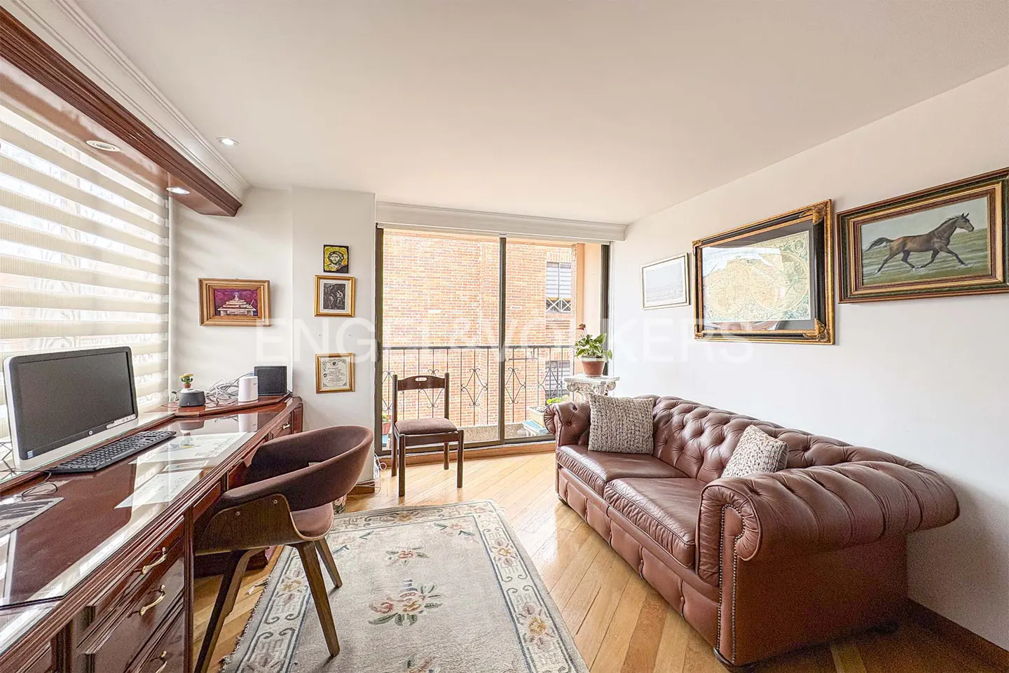 A living room with a brown leather sofa, wooden floors, and a desk with a computer. Balcony in background.