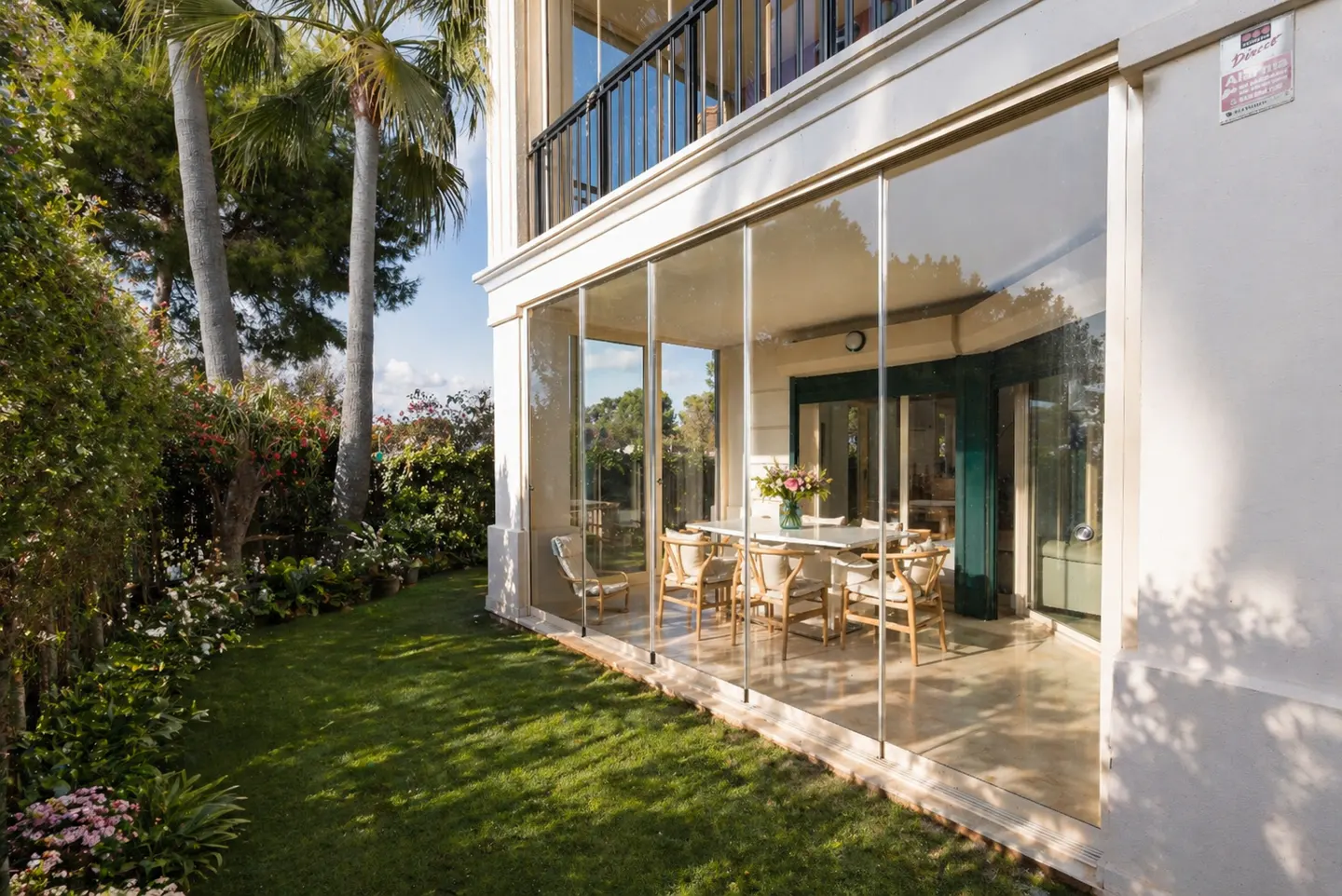 Exterior view of a home with a glass-enclosed patio, table, chairs, and a green lawn with trees.