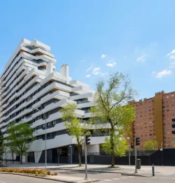 Modern white apartment building with tiered balconies next to a traditional brick building under a clear blue sky.