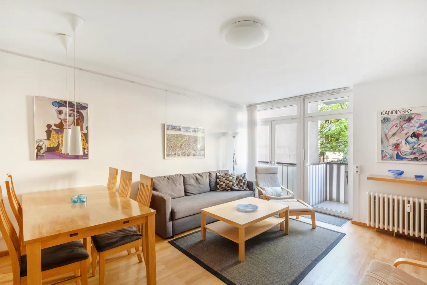 Bright living room with wood floors, gray sofa, wood table and chairs, and balcony access. Art adorns the white walls.