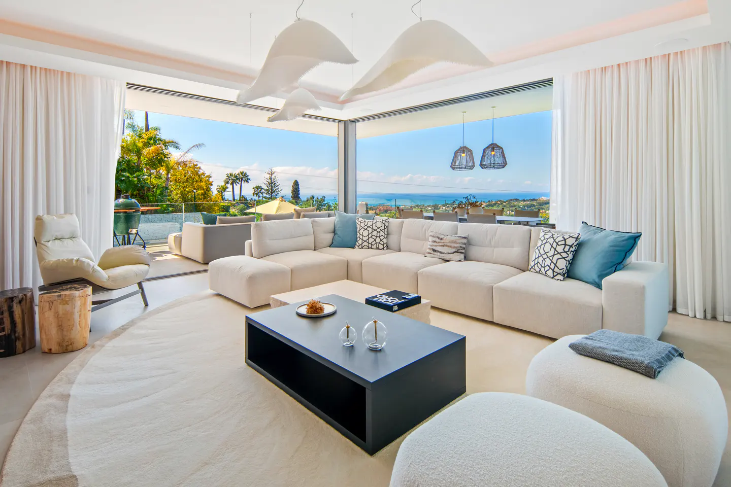 Bright living room with white sectional sofa, black coffee table, and ocean view through large windows. White curtains and modern lighting fixtures.