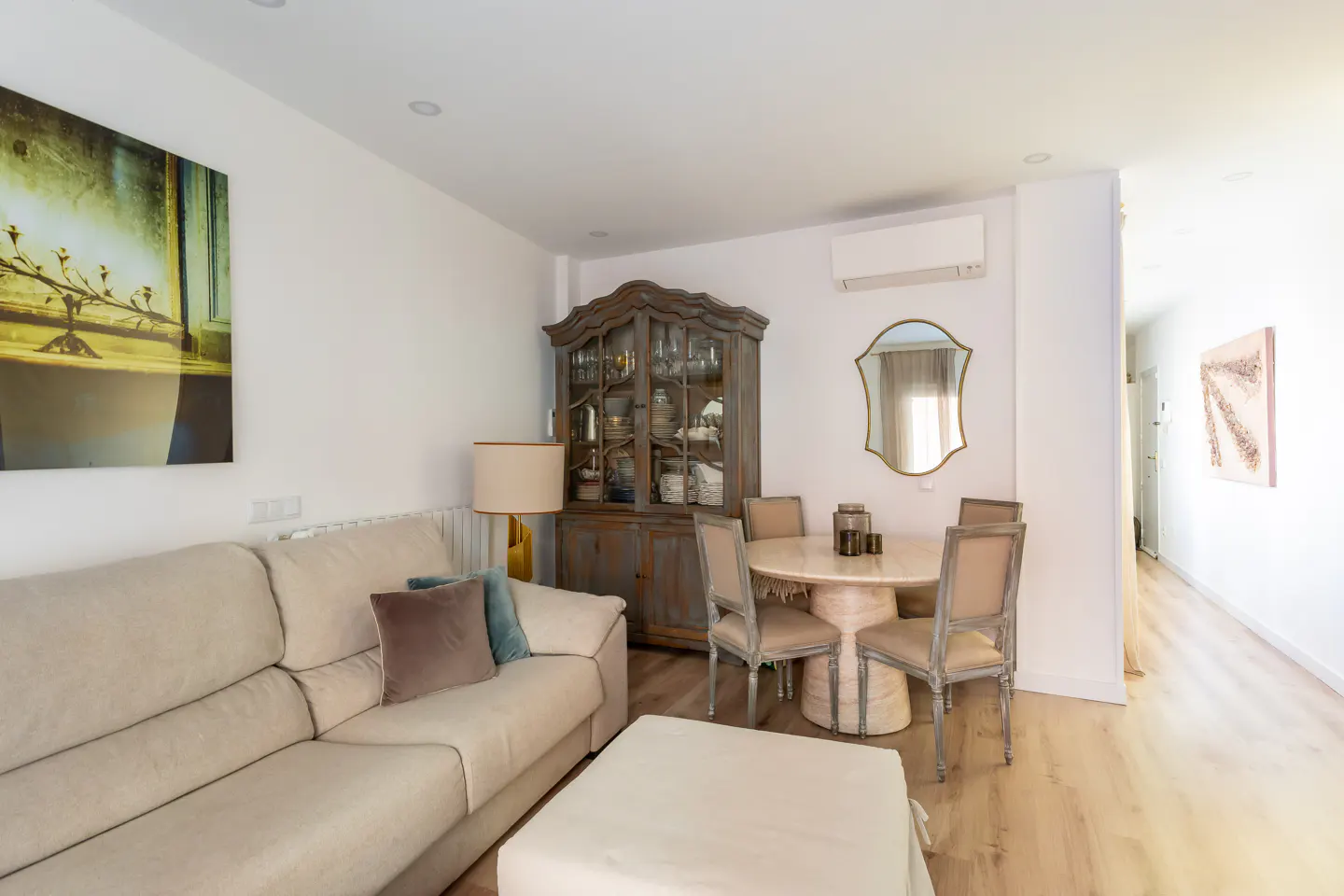 Living room with a beige sofa, round table with four chairs, and a wooden china cabinet against a white wall.