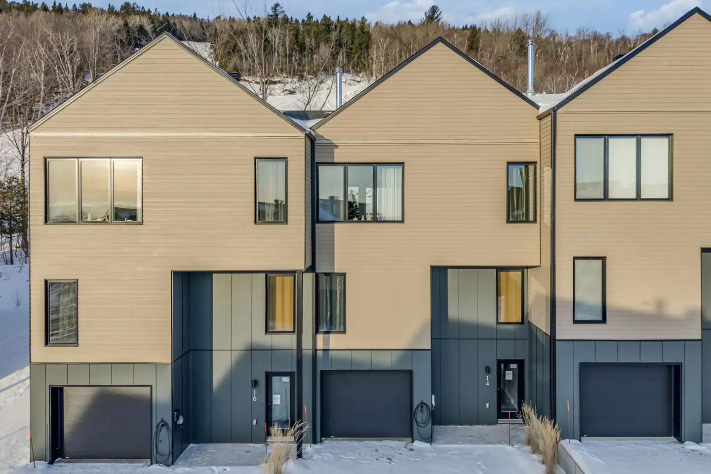 Three modern townhouses with beige siding and dark gray accents stand in a snowy landscape. Each has a garage and multiple windows.