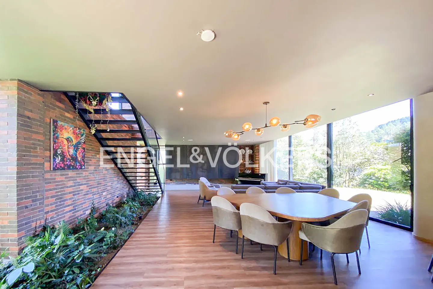 Open-concept living space with a dining table, stairs, and a large window with a view of greenery. Brick wall and wood floors.