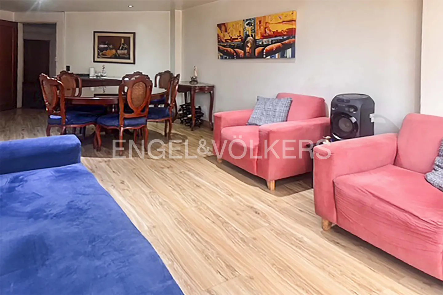Living room with blue sofa, pink armchairs, and wood floor. A dining table with chairs is in the background.