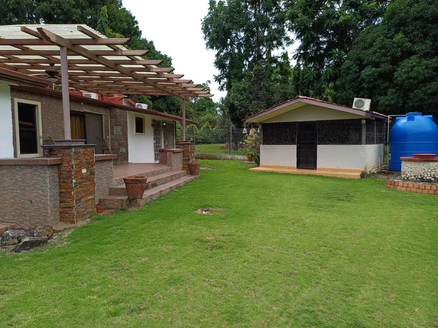 Exterior view of a house with a pergola, a small building, and a blue water tank on a green lawn.