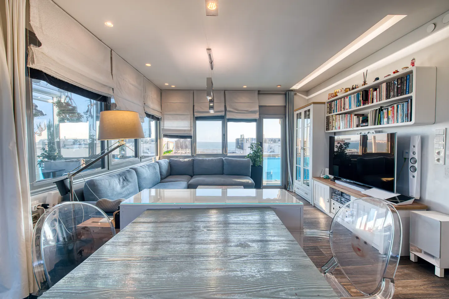 Bright living room with a gray sectional sofa, a wooden table with clear chairs, and a TV with a bookshelf. Ocean view through the windows.