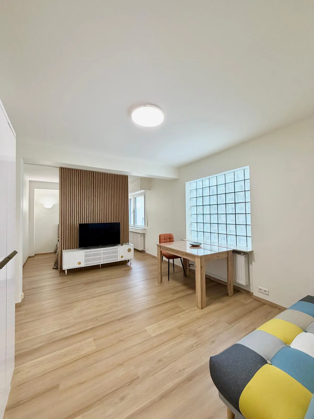 Modern living room with light wood floors, a slatted wood accent wall, a TV console, dining table, and colorful sofa.