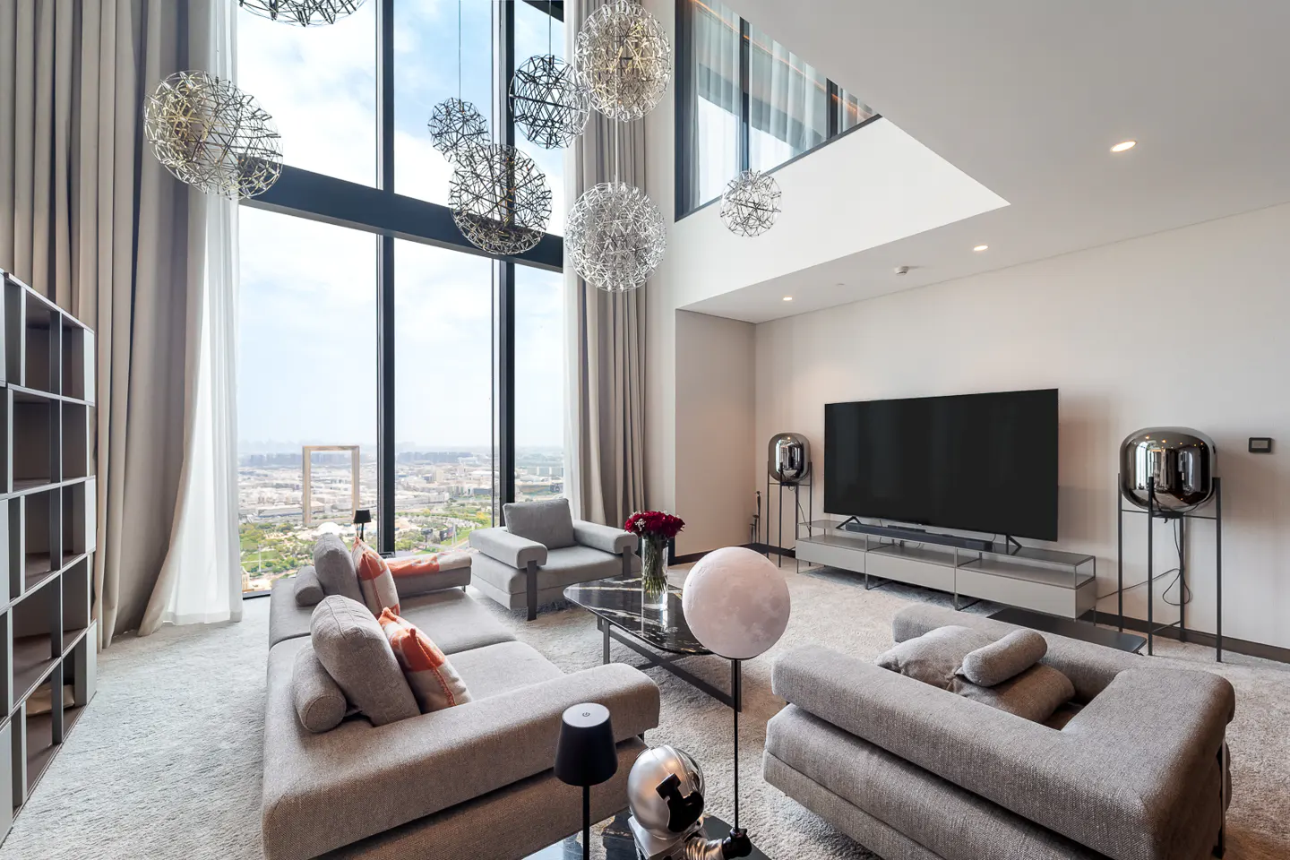 A modern living room with gray sofas, a large TV, and floor-to-ceiling windows overlooking a city. Decorative chandeliers hang from the high ceiling.