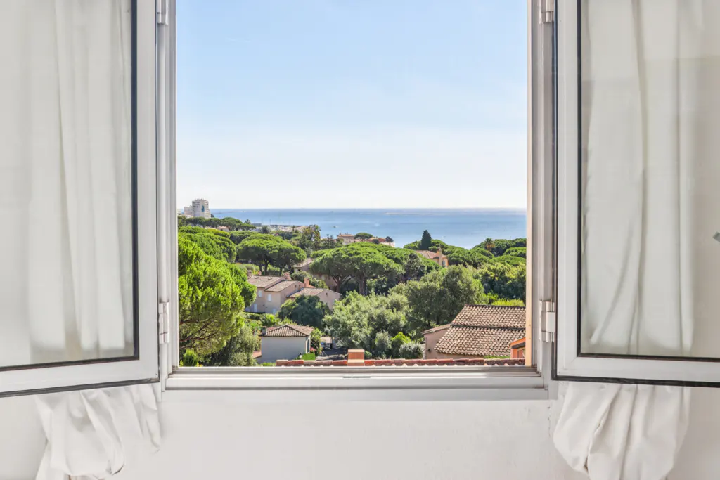 View from an open window with white curtains, overlooking green trees, houses, and the blue ocean under a clear sky.