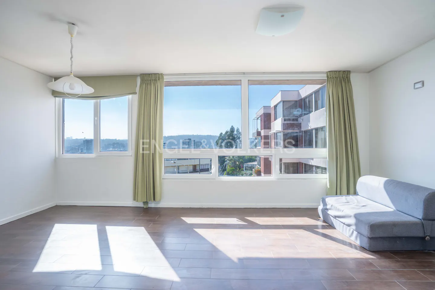 Bright, empty room with hardwood floors, white walls, and large windows with green curtains. A light blue sofa sits against the wall.