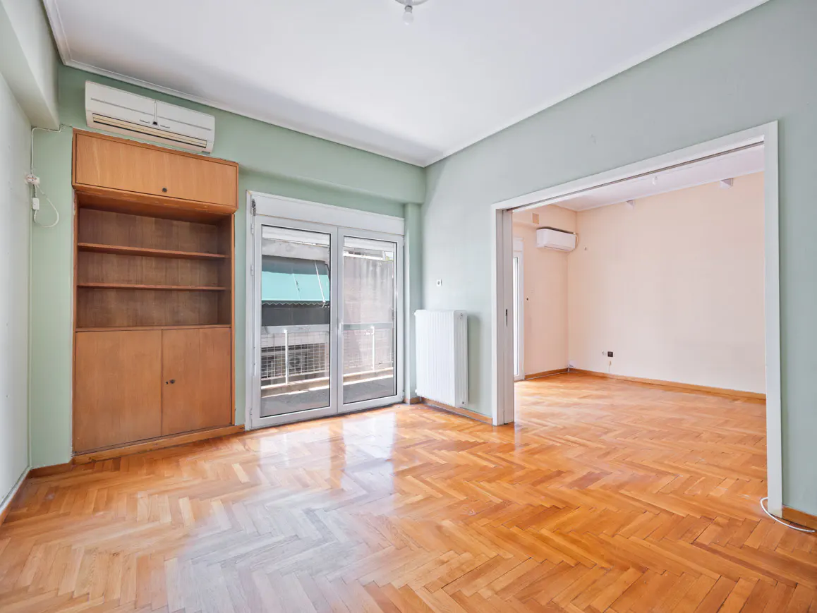 Empty living room with light green walls, herringbone wood floors, a wooden built-in shelf, and a doorway to another room.