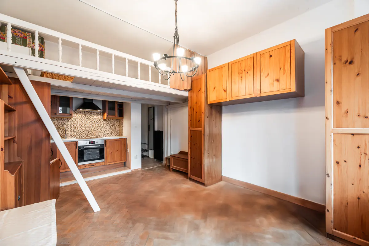 Cozy apartment interior with wood floors, cabinets, and a loft. A small kitchen is visible with mosaic backsplash and stainless steel oven.