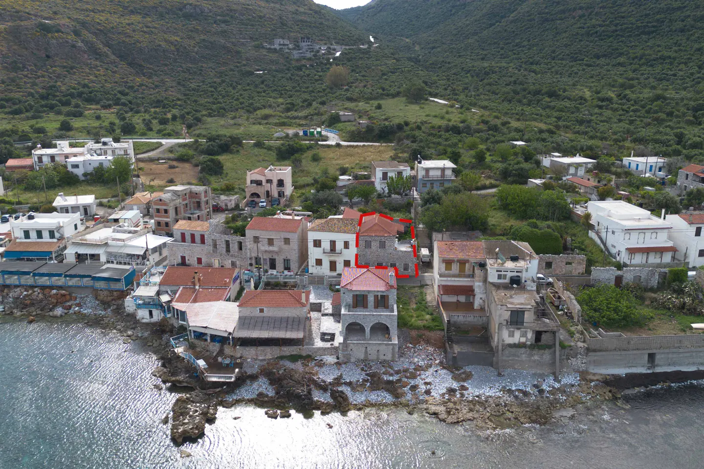 Aerial view of a coastal village with stone houses and a red-outlined property. Green hills rise in the background.