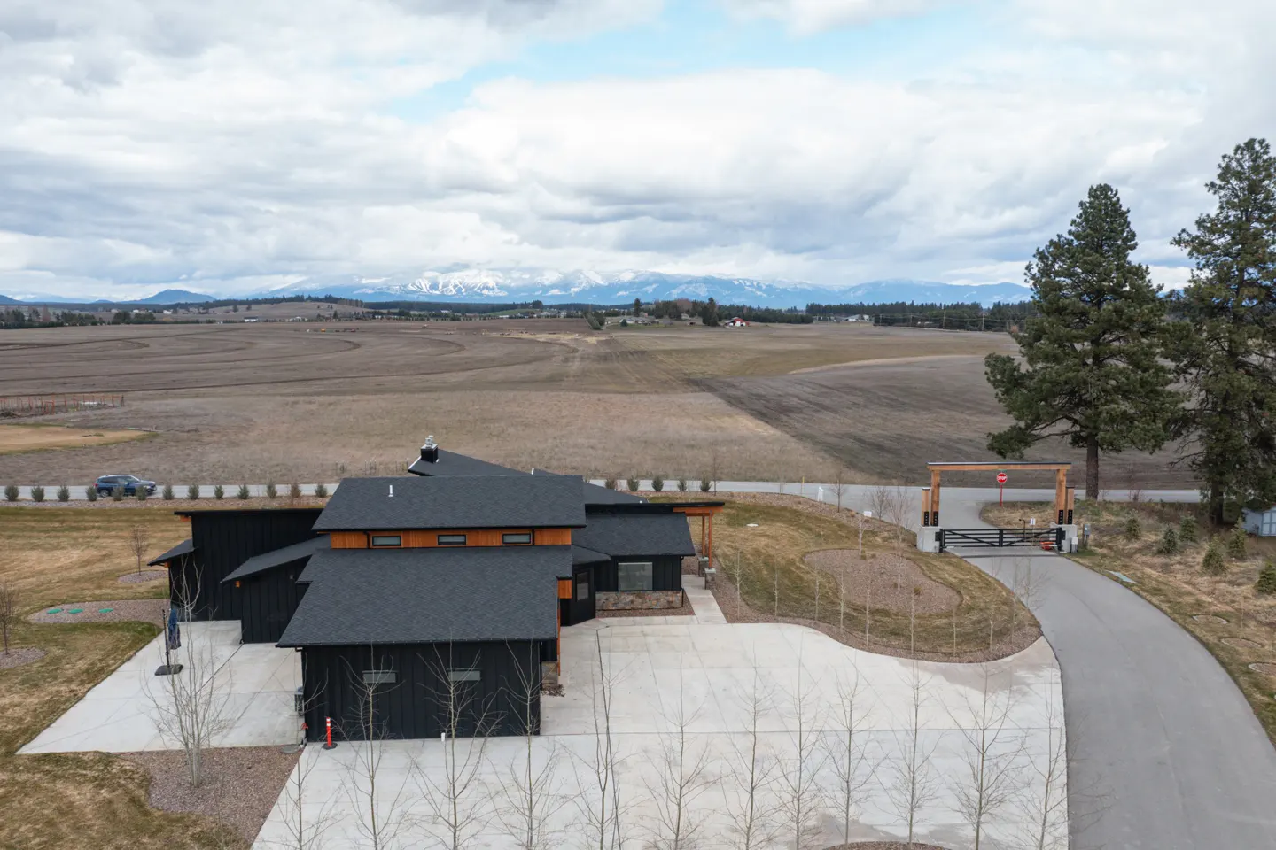 Aerial view of a modern black house with a gray roof, a long driveway, and a large field with mountains in the background.