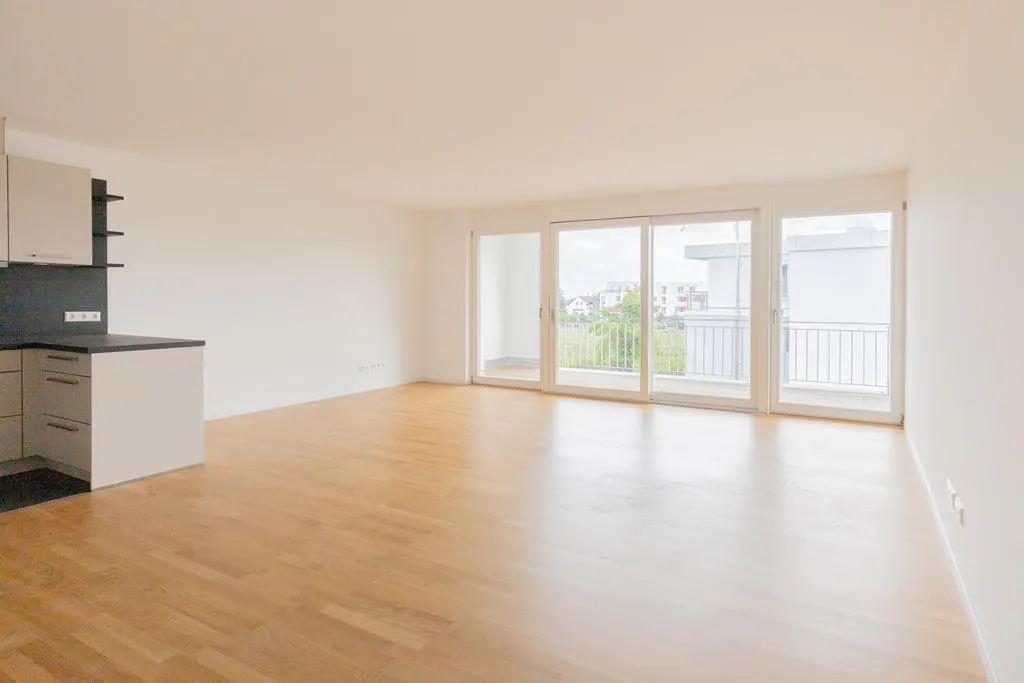 Bright, empty apartment with wood floors, white walls, and a kitchen area. Sliding glass doors lead to a balcony.