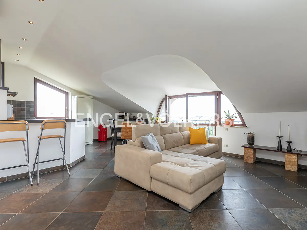 Attic apartment with a beige sectional sofa, yellow pillow, and dark tile floors. Kitchen bar with stools and windows with a view.