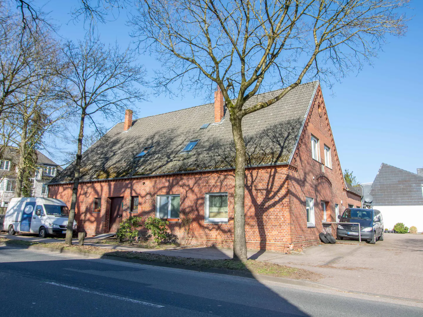 Two-story red brick house with a gray roof, chimneys, and bare trees in the front yard on a sunny day.