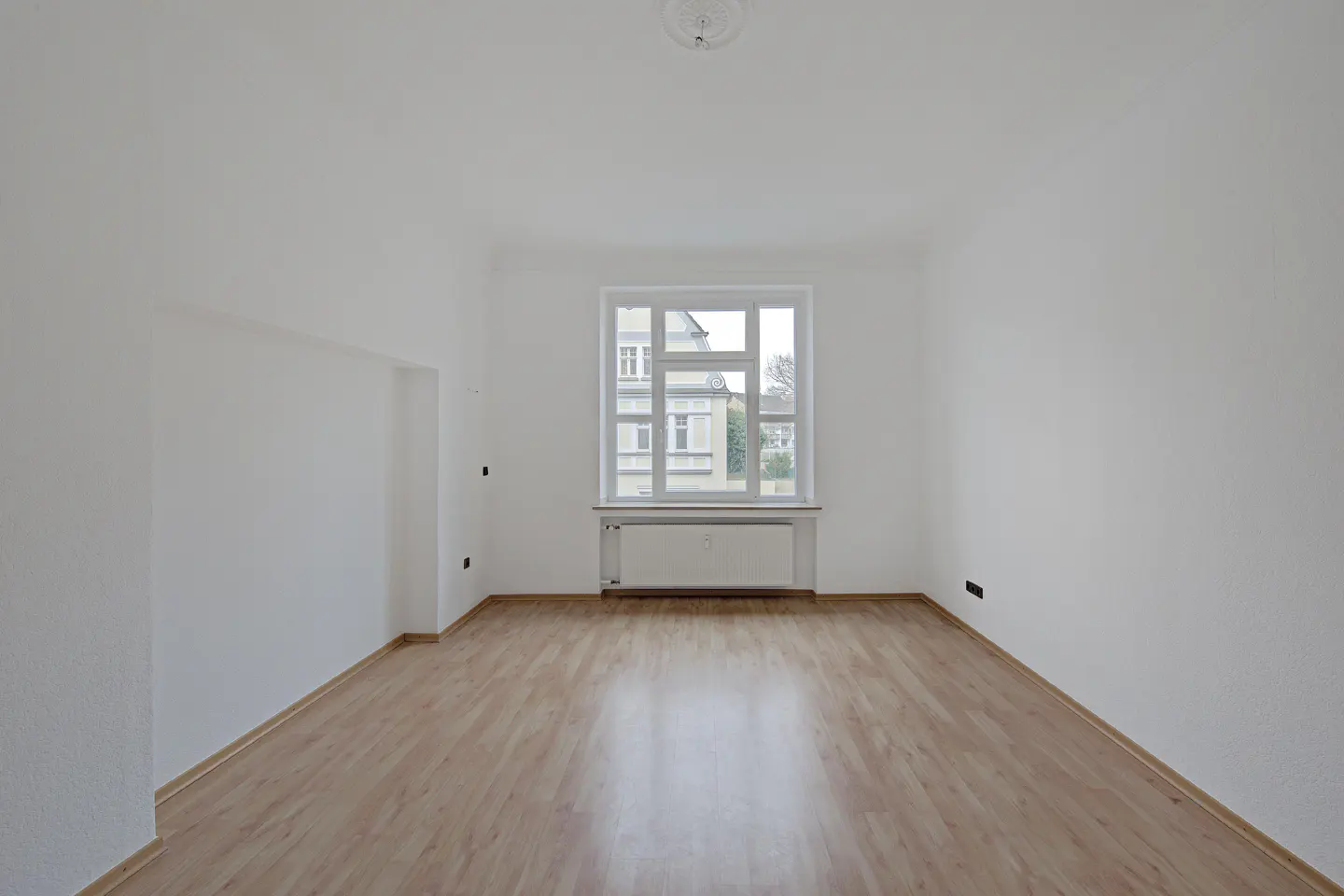 Empty room with light wood floors, white walls, and a large window. A radiator sits beneath the window. Outside, a building is visible.