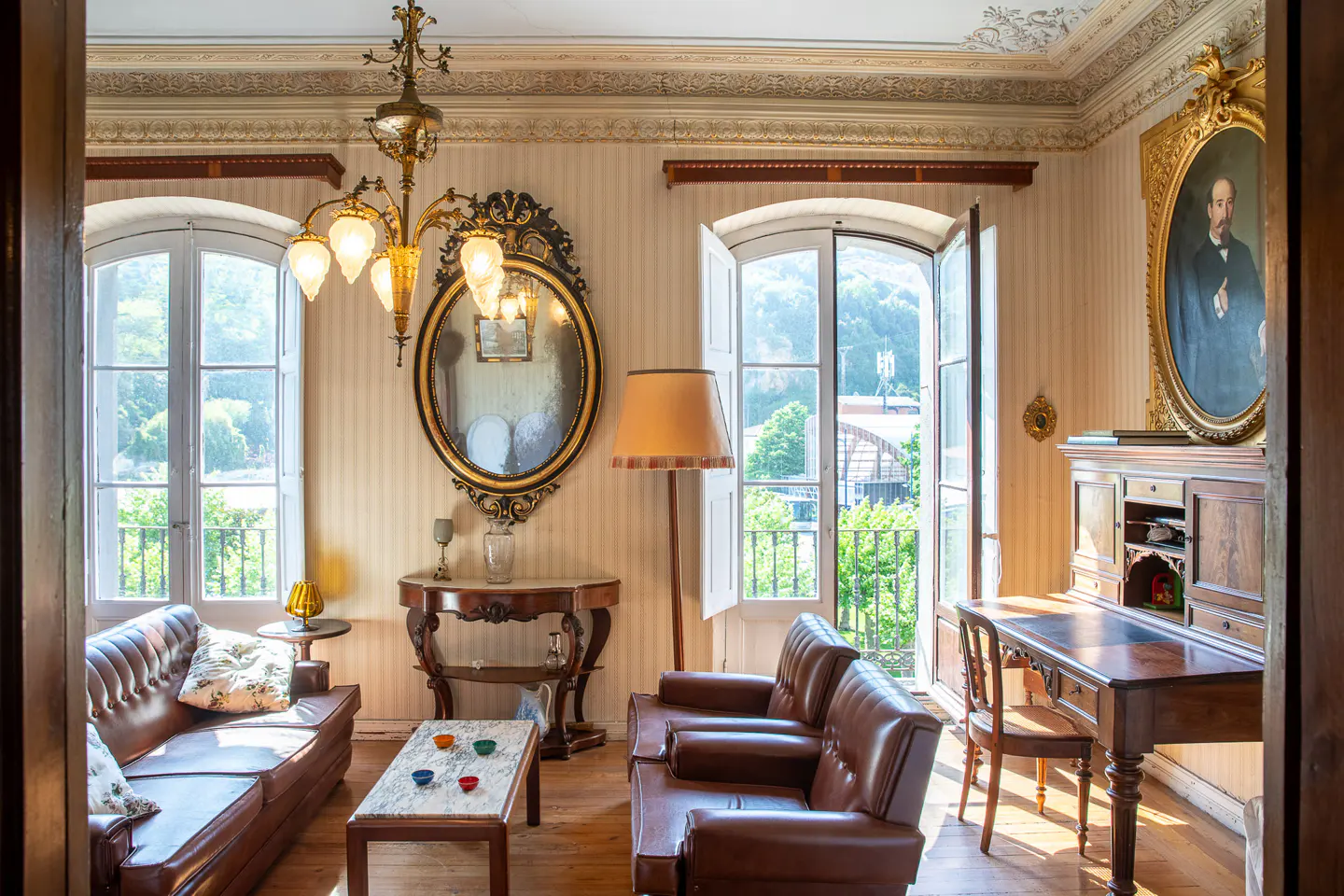 Ornate living room with brown leather sofas, marble-topped tables, and a vintage desk. Large windows offer a view of lush greenery.