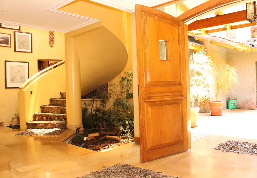 Open wooden door reveals a yellow-walled foyer with a spiral staircase, plants, and marble floors. Pictures hang on the wall.