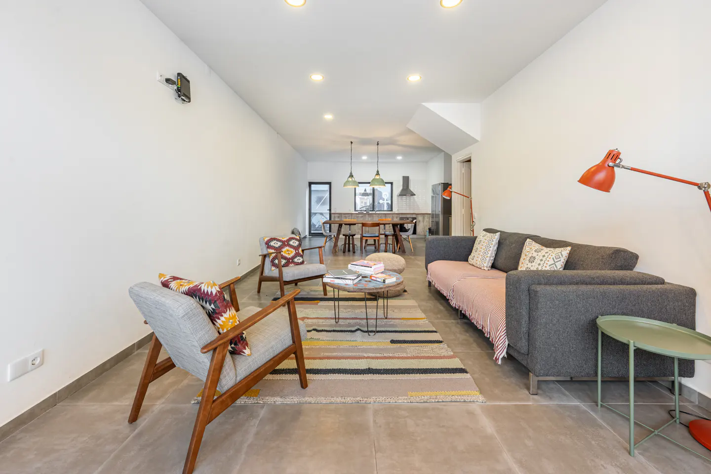 Bright, modern living room with gray sofa, wooden chairs, and colorful rug. Kitchen and dining area visible in the background.