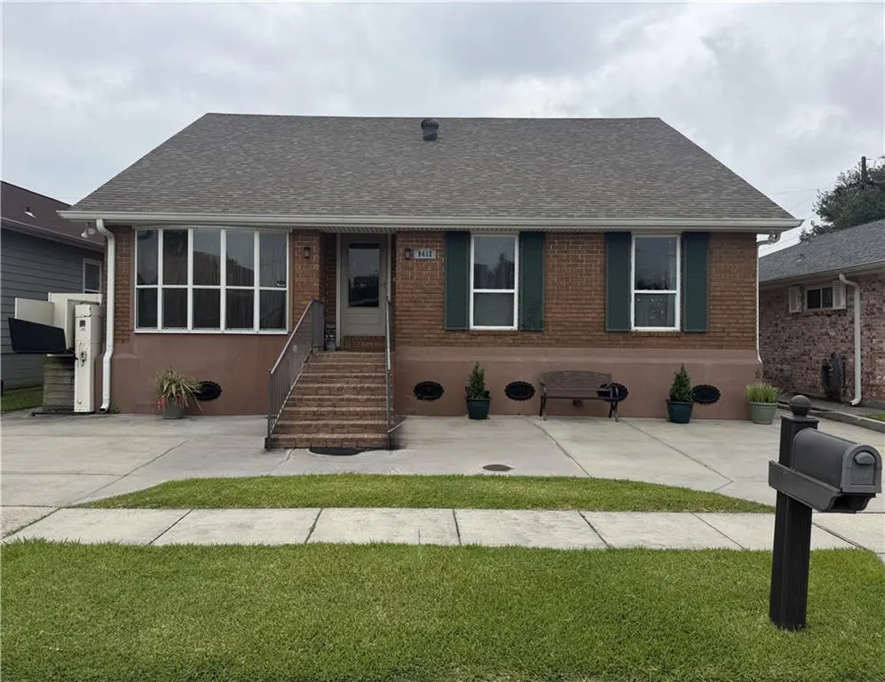 A single-story brick house with a gray roof, green shutters, and a concrete driveway. A mailbox stands on the lawn.