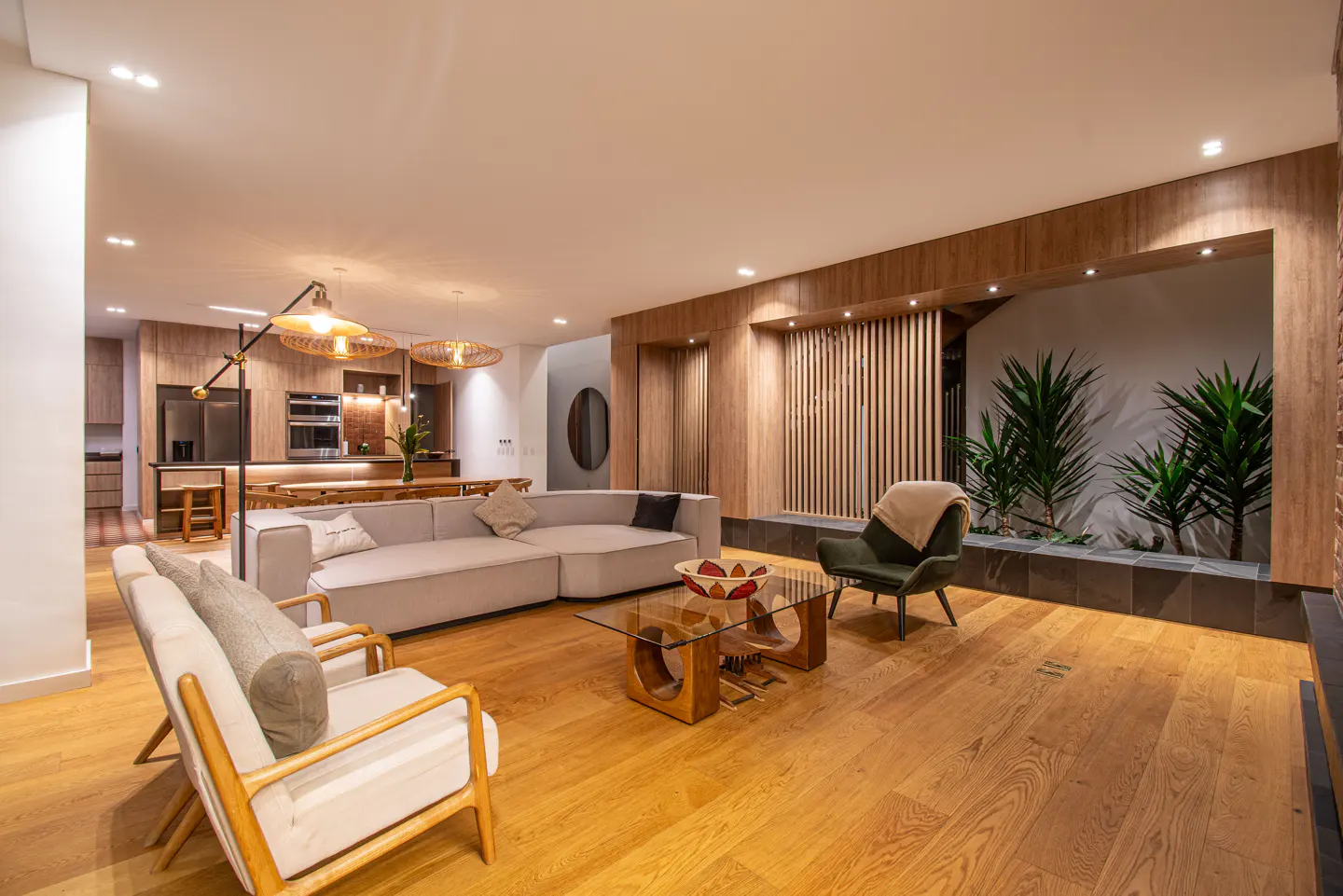 Open-concept living room with wood floors, a gray sectional sofa, and a green armchair. A glass coffee table sits in the center. A kitchen is visible in the background.
