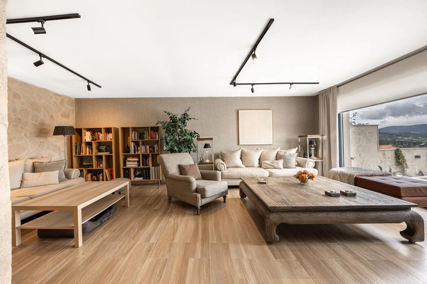 Bright living room with wood floors, stone wall, and large windows. Sofas, chairs, and bookshelves furnish the space.