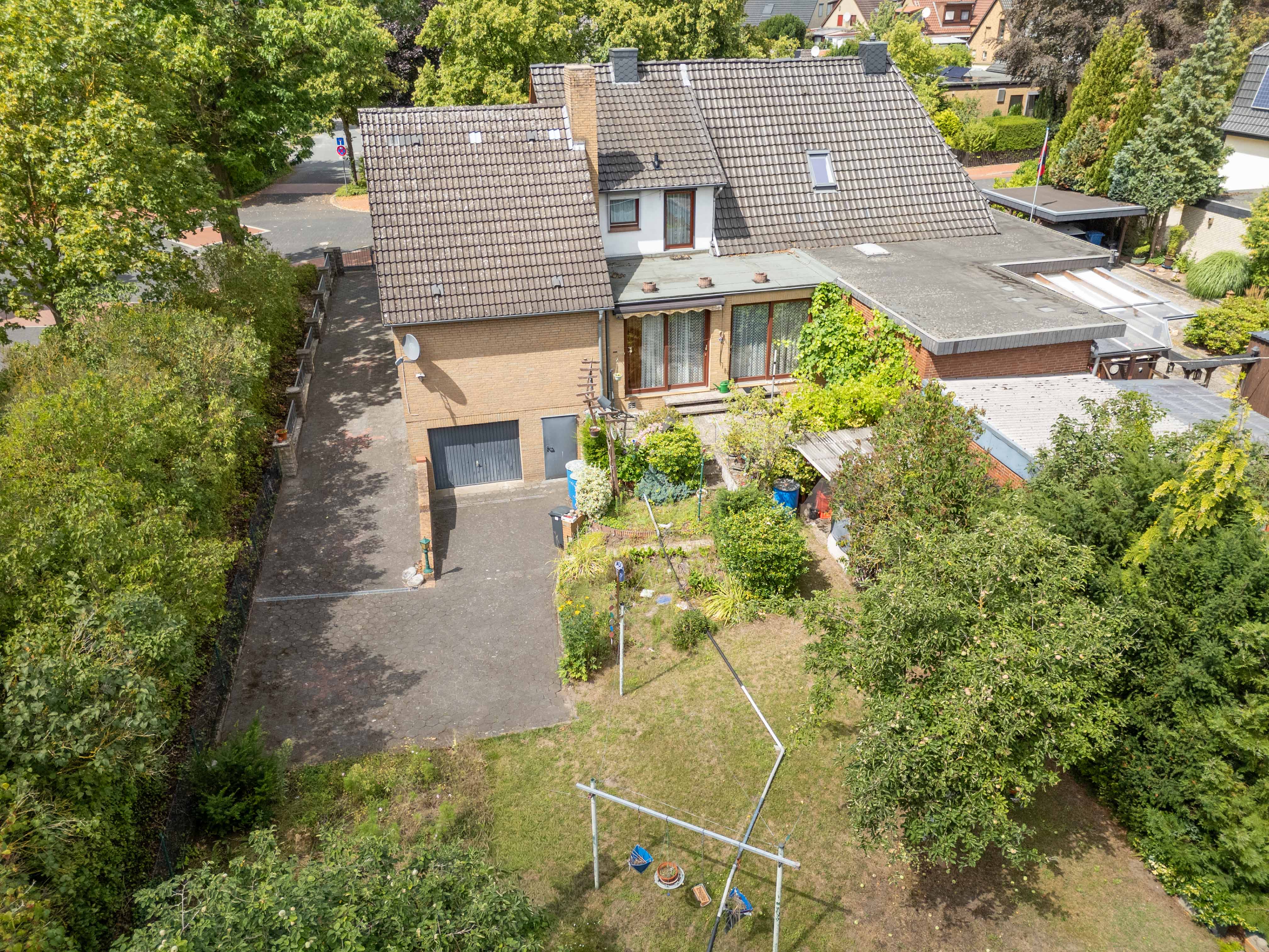 Aerial view of a tan brick house with a tiled roof, a paved driveway, and a large, tree-filled backyard with a clothesline. [AI-generated text]