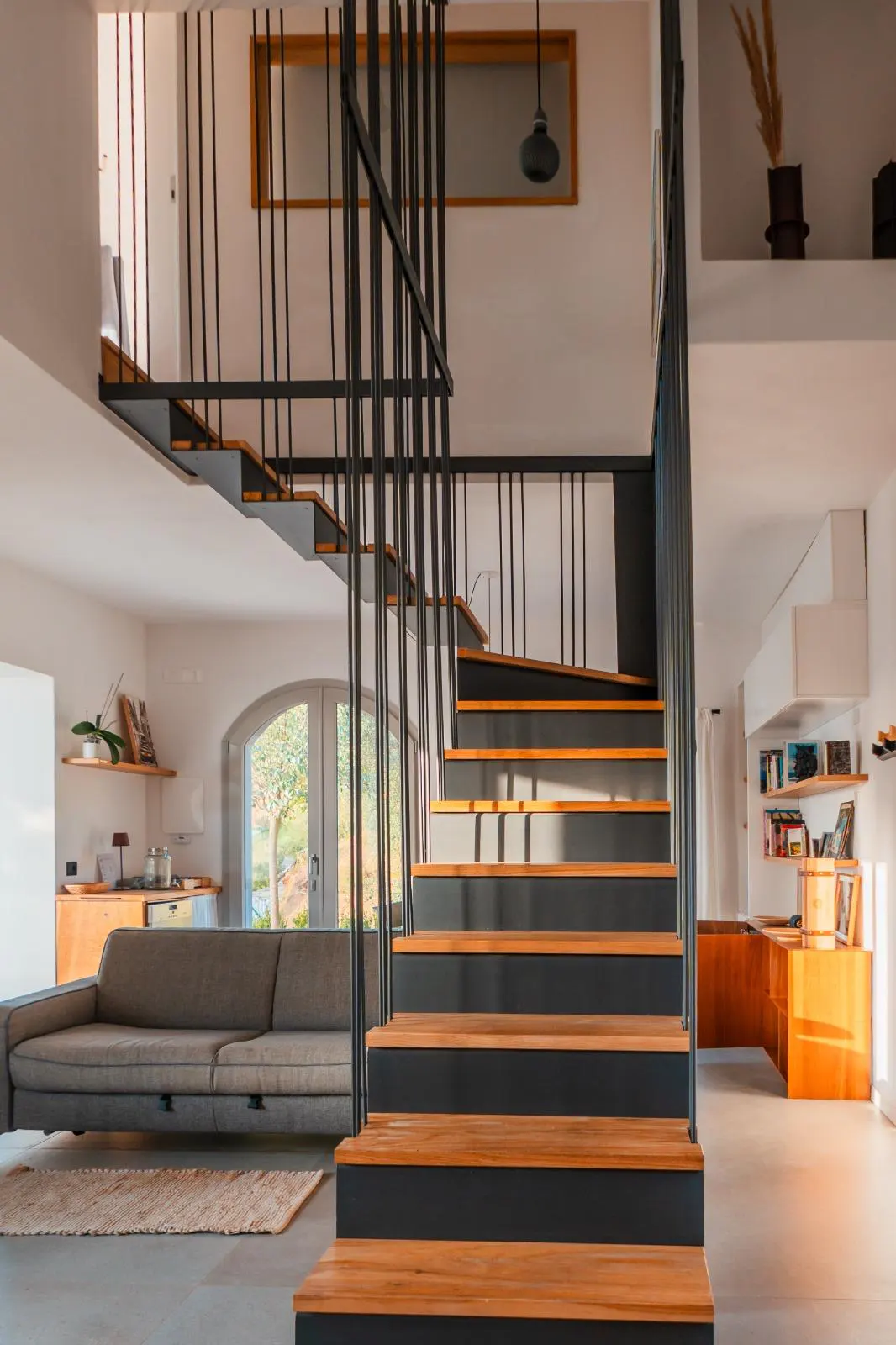 Interior view of a modern home featuring wooden stairs with black railings, a gray sofa, and arched glass doors.