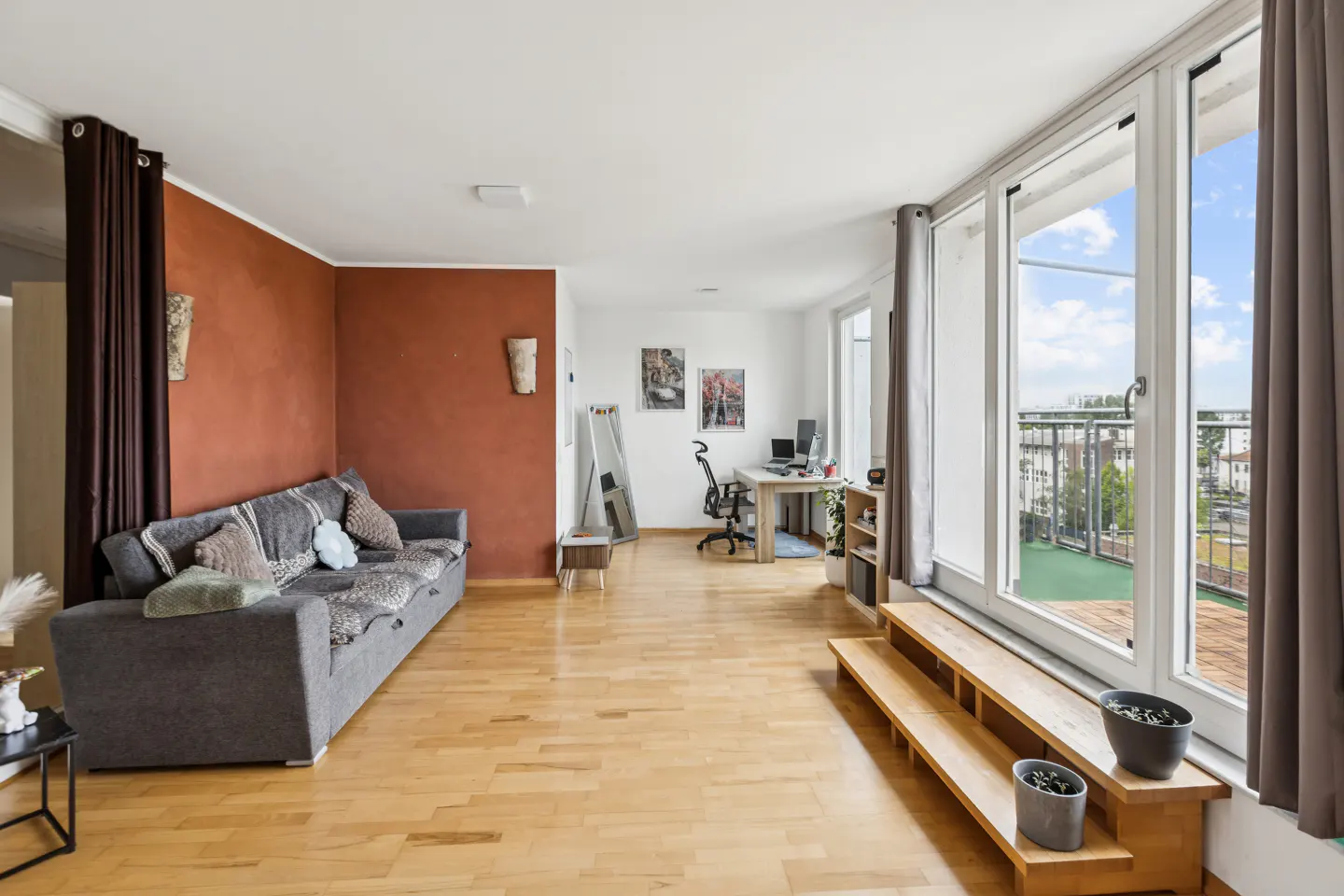 Living room with hardwood floors, a grey sofa against a terracotta wall, a home office area, and large windows to a balcony.